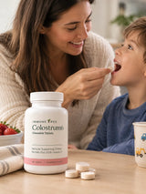 Immune Tree Colostrum6 Strawberry Moo Chews lifestyle image showing a smiling mother giving a chewable colostrum tablet to her young son at a kitchen table, with the Colostrum6 bottle and loose tablets in the foreground, a bowl of fresh strawberries to the left, and the child's cup to the right